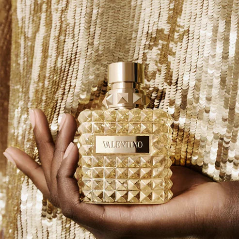 Gold Valentino perfume bottle held by a hand against a gold sequin background