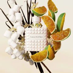 Valentino perfume bottle surrounded by vanilla beans, oranges, and flowers on a beige background